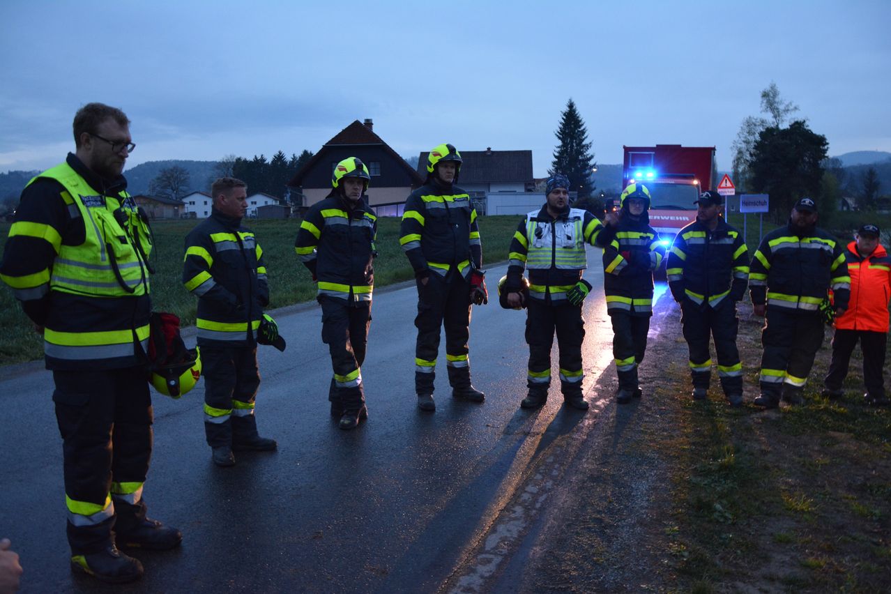 Fünf Feuerwehrleute stehen auf einer Straße, tragen reflektierende Anzüge und Helme, möglicherweise im Dienst. Hinter ihnen steht ein roter Feuerwehrwagen mit eingeschalteten Lichtern. In der Ferne sind Häuser und Bäume zu sehen.