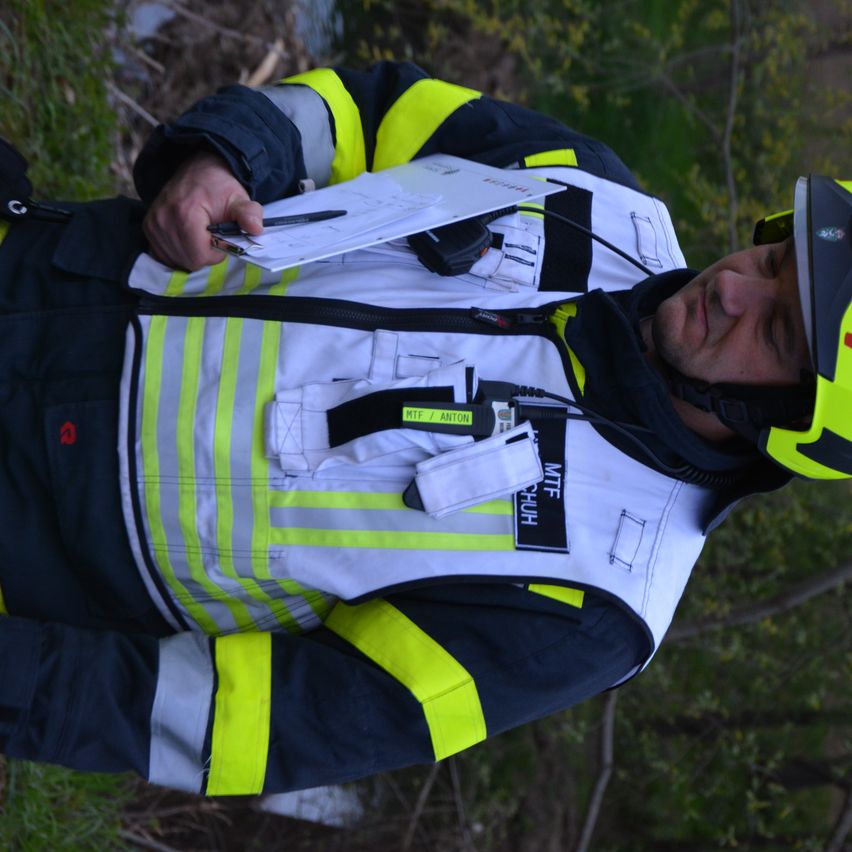 Ein Feuerwehrmann ruht auf Gras mit einem Helm, hält Papier und Stift. Er trägt eine reflektierende Weste.