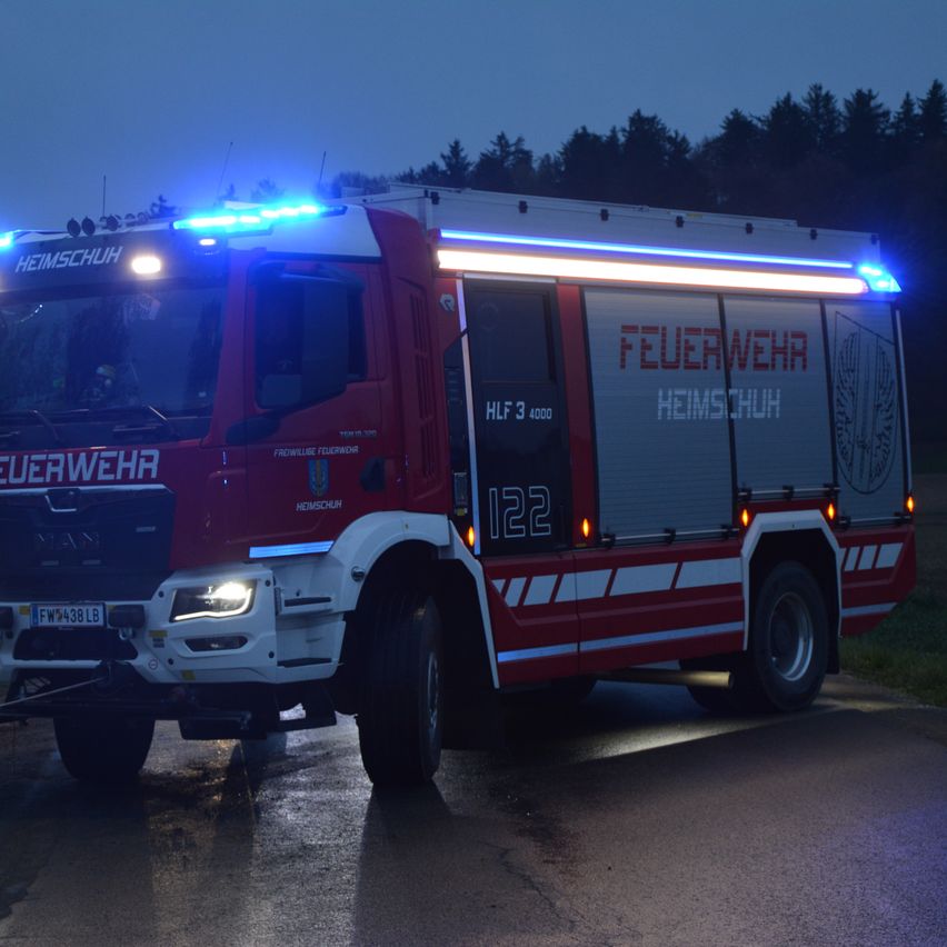 Ein roter Feuerwehrwagen mit blauen Lichtern steht nachts auf einer Straße. Er hat die Aufschrift 'FEUERWEHR' und 'HEIMSCHUH' an der Seite. Die Front hat 'FW:498LB'. Bäume sind dahinter.