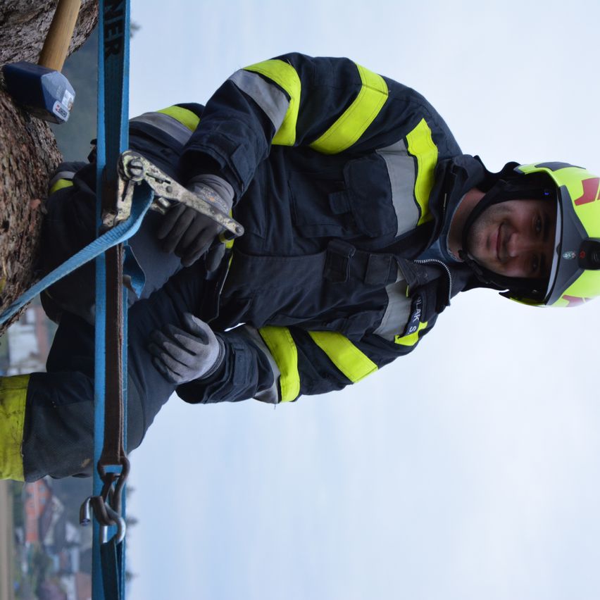 Ein Feuerwehrmann klettert mit Sicherheitsseilen und einem Gurtzeug an einem Baum hoch, trägt einen gelben und schwarzen Overall und Helm. Er lächelt und scheint für ein Foto zu posieren.