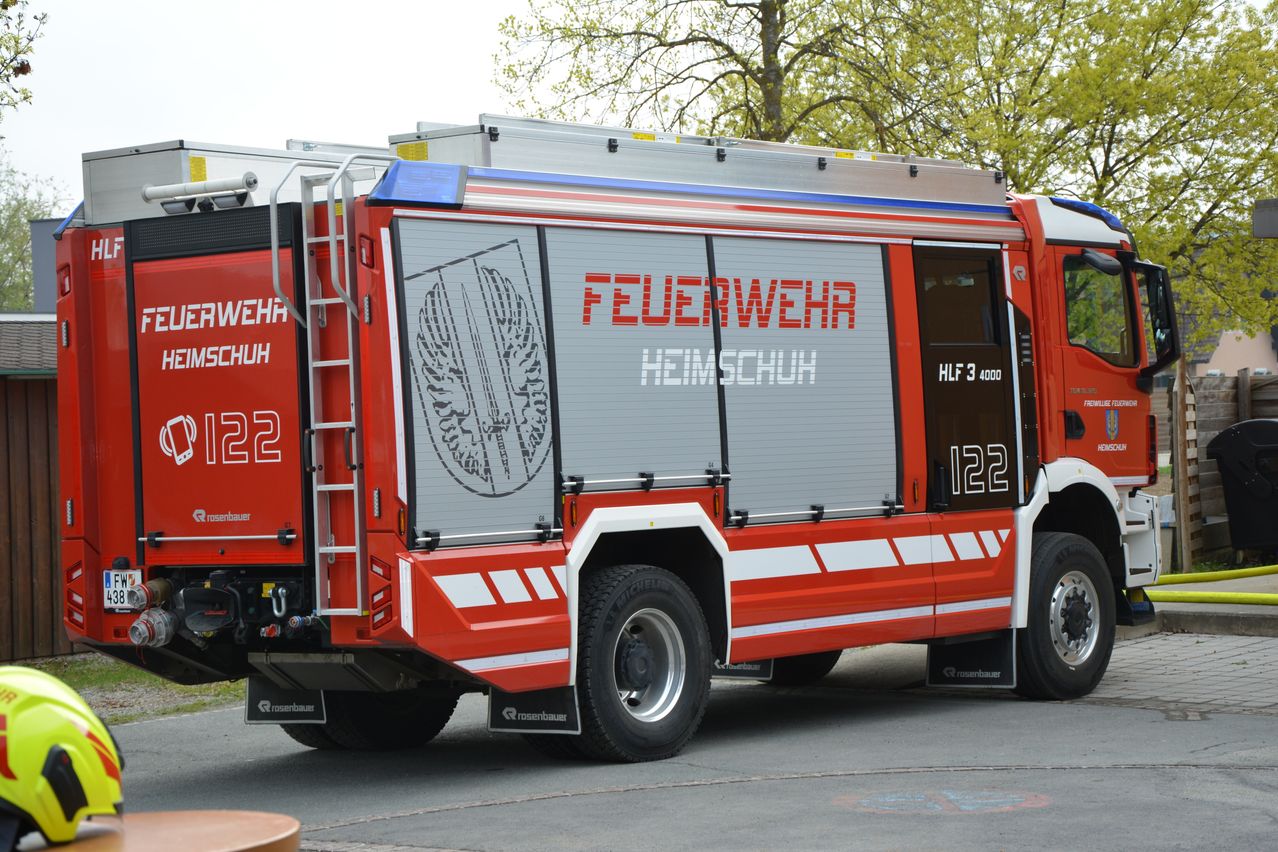 Die Rückseite eines roten Feuerwehrwagens aus Heimschuh, Österreich, zeigt den Text 'Feuerwehr Heimschuh'. Der Wagen ist auf einem gepflasterten Bereich geparkt, mit Bäumen im Hintergrund.
