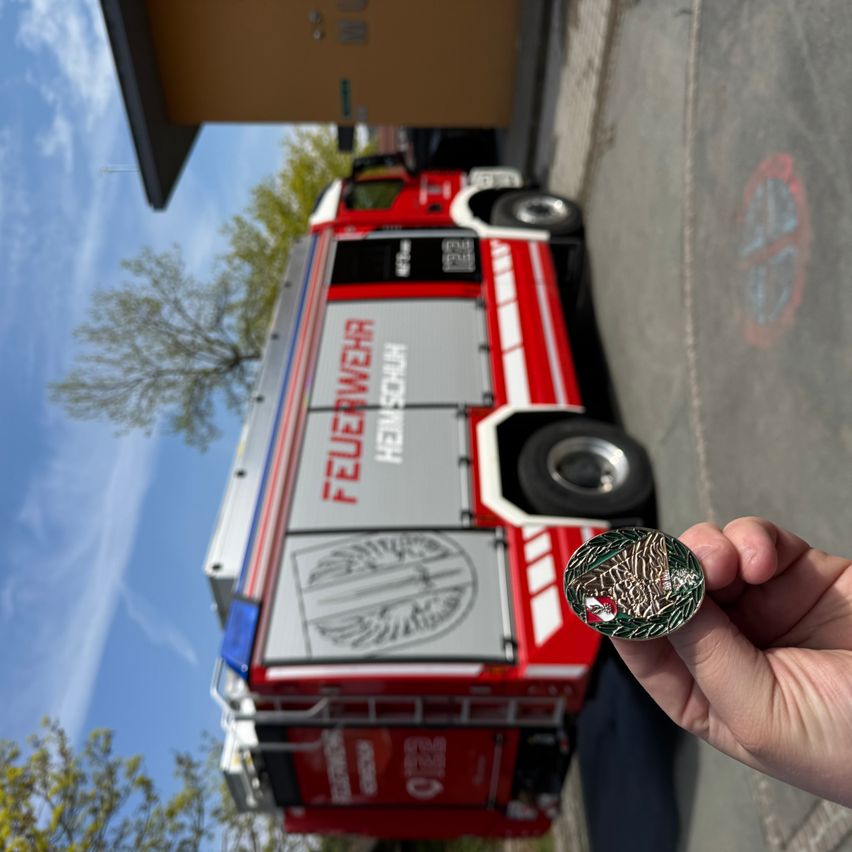 Ein Feuerwehrfahrzeug ist am Straßenrand geparkt, und eine Person hält eine Münze mit einem Feuerwehremblem.
