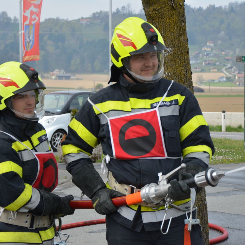 Zwei Feuerwehrleute in voller Ausrüstung stehen neben einem Baum, einer hält eine Feuerwehrspritze. Ein Auto und eine Fahne im Hintergrund.