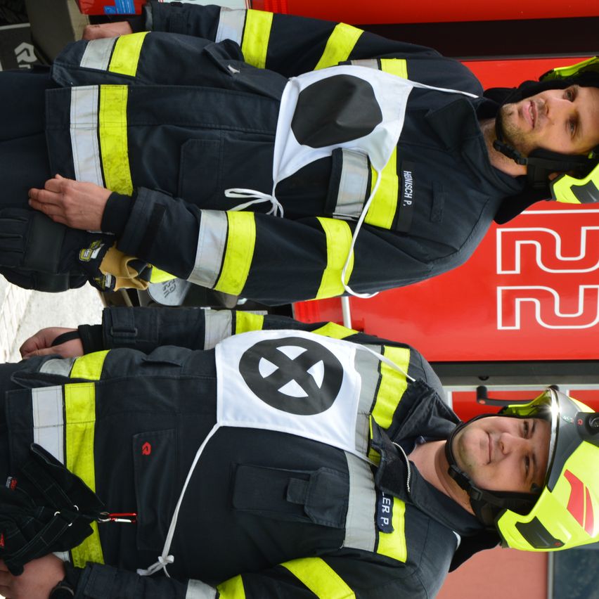Zwei Feuerwehrleute stehen nebeneinander vor einer Feuerwache. Beide tragen Helme und Uniformen. Der vordere hat ein rotes Abzeichen auf der Schulter.