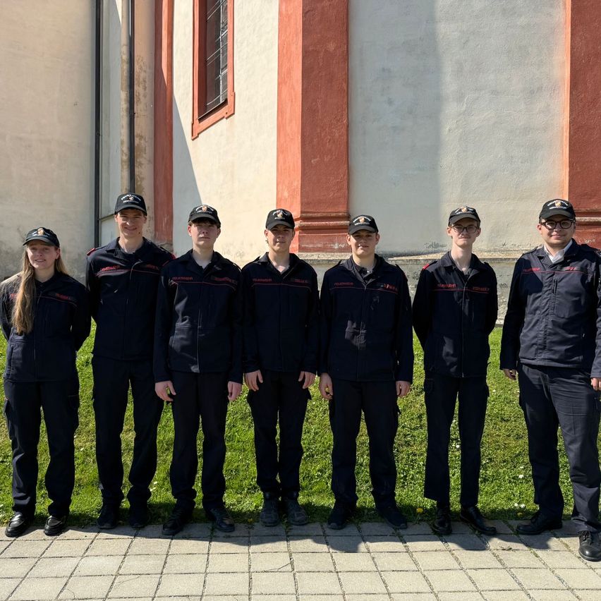Eine Gruppe von neun Personen in schwarzen Uniformen steht vor einem Gebäude mit roten Säulen und einem Fenster. Sie posieren wahrscheinlich für ein Foto.