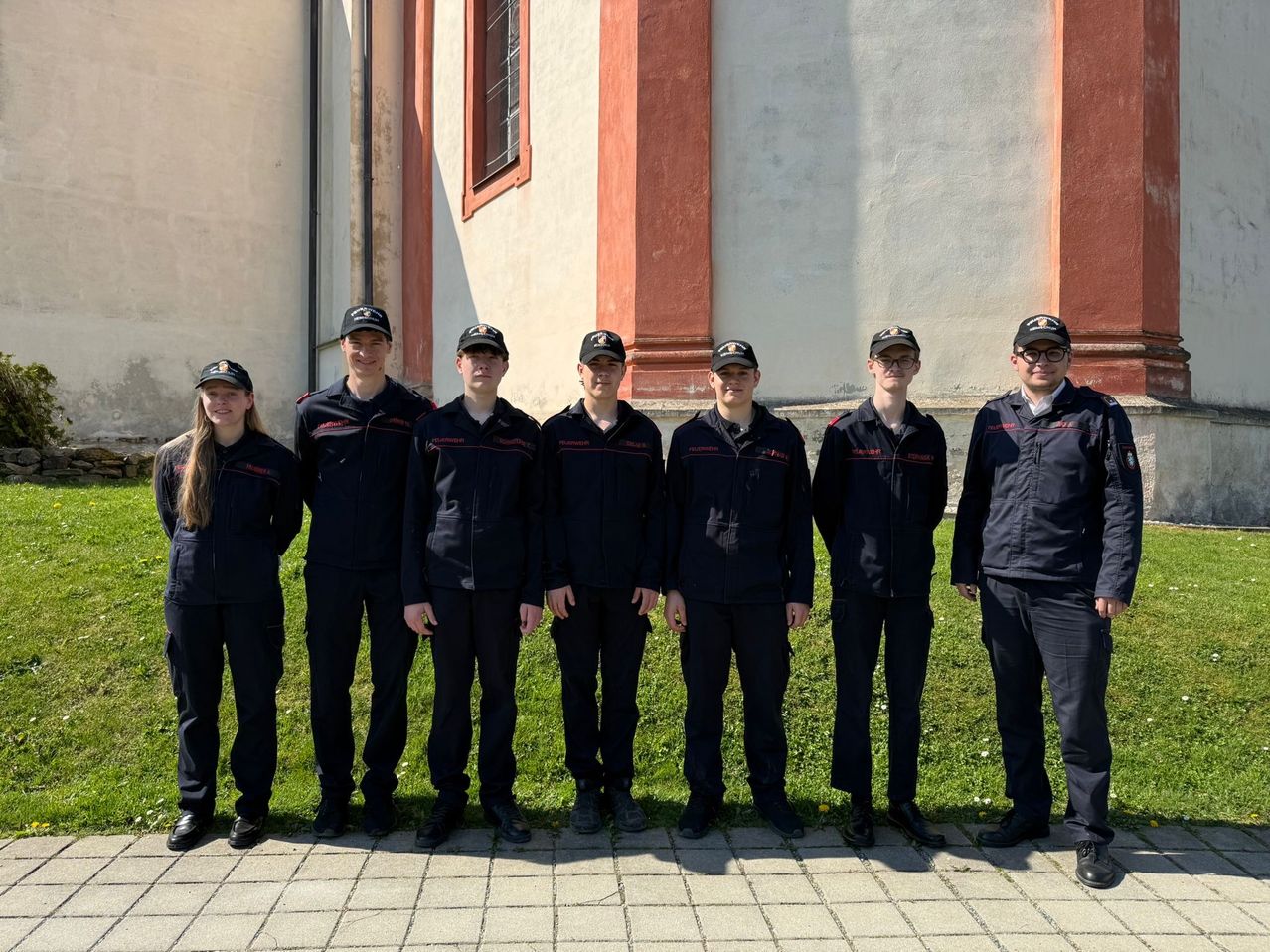 Eine Gruppe von neun Personen in schwarzen Uniformen steht vor einem Gebäude mit roten Säulen und einem Fenster. Sie posieren wahrscheinlich für ein Foto.