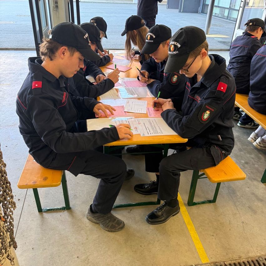 Eine Gruppe junger Menschen in Uniformen sitzt um einen Tisch und schreibt auf Papiere. Sie sind drinnen, wahrscheinlich in einer Trainingseinrichtung.
