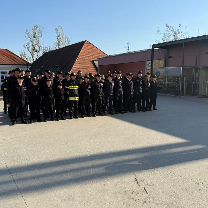 Eine Gruppe Feuerwehrleute in Uniform steht in Formation vor einigen Gebäuden unter klarem Himmel.