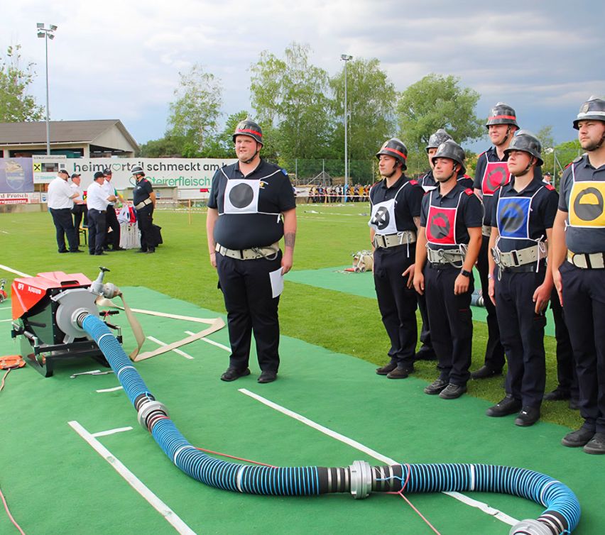 Eine Gruppe von Männern in Uniformen steht auf einem Feld mit Schutzausrüstung. Ein Mann in der Mitte hält ein Papier, und andere beobachten. Eine Maschine mit einem Schlauch liegt in der Nähe.