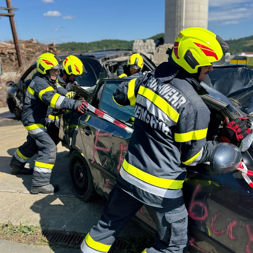 Drei Feuerwehrleute in gelben Helmen und Uniformen extrahieren mit Werkzeugen eine Person aus einem beschädigten Auto. Das Auto ist demoliert, und die Feuerwehrleute sind am Straßenrand.