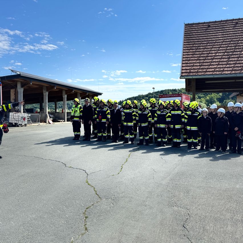 Eine Gruppe Feuerwehrleute steht in Formation im Freien, trägt Helme und Uniformen unter einem klaren Himmel. In der Nähe sind ein Gebäude und ein Feuerwehrwagen zu sehen.
