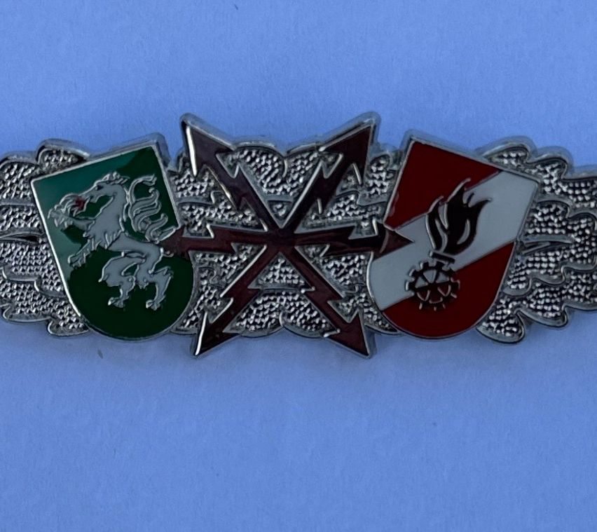 Eine silberne Brosche mit drei Emblemen. Links ist ein grüner Schild mit einem weißen Löwen, in der Mitte ein roter Kreuz mit Pfeilen und rechts ein rot-weißer Schild mit einer Flamme.