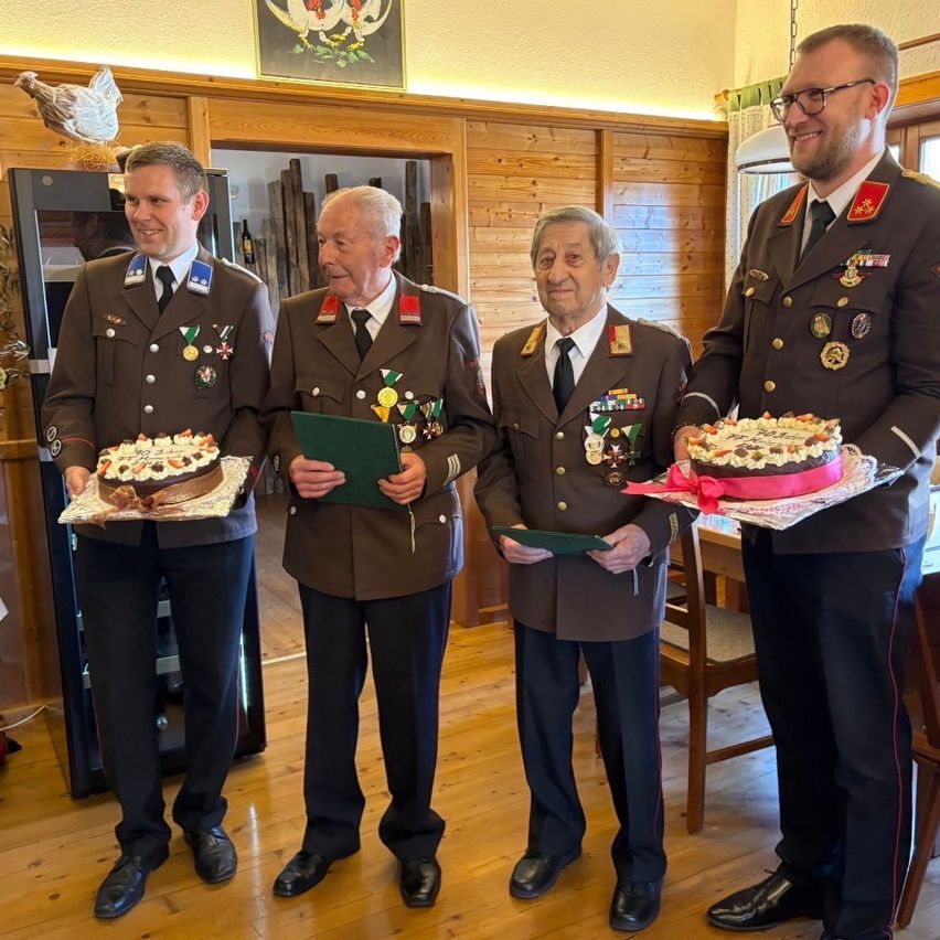 Vier Männer in Militäruniformen stehen zusammen und lächeln. Sie halten dekorierte Kuchen und eine grüne Mappe. Hinter ihnen ist eine Holzwand mit einem Bilderrahmen sichtbar.