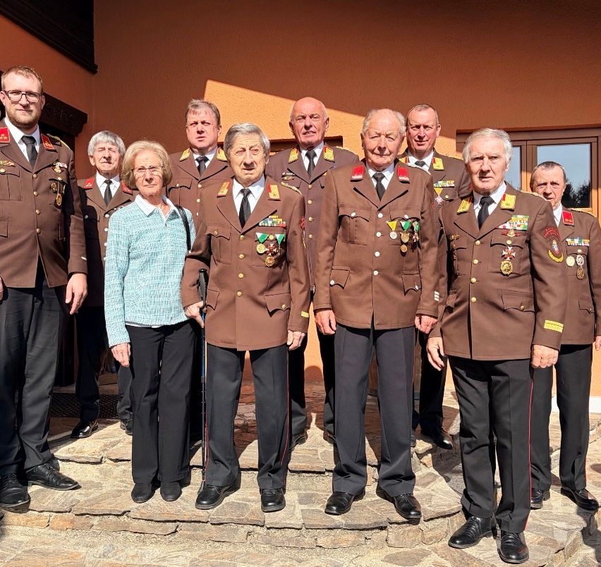 Eine Gruppe älterer Personen in Militäruniformen steht zusammen, mit einer Frau in einem hellblauen Pullover in der Mitte. Sie posieren für ein Foto auf Steinstufen.