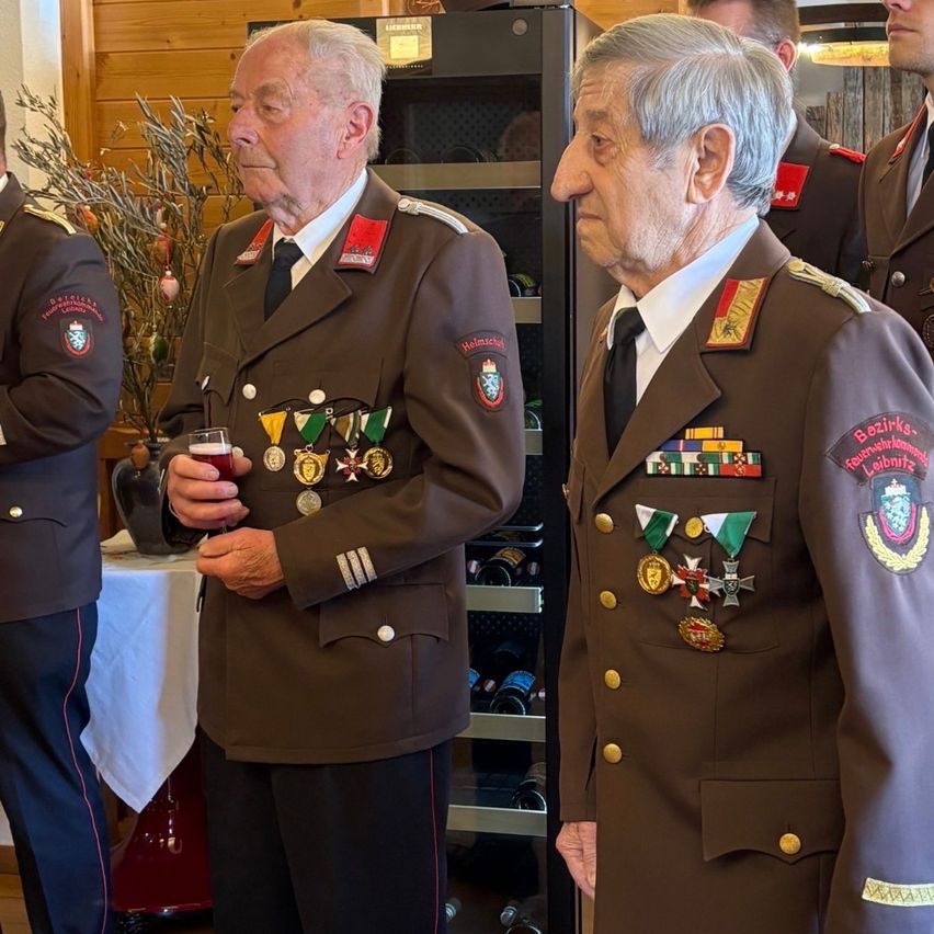 Mehrere Männer in Militäruniformen stehen zusammen; einer trinkt ein Getränk, während andere nach vorne schauen. Sie tragen Medaillen an ihren Uniformen. Hinter ihnen ist eine Holzwand und ein Regal mit Flaschen.