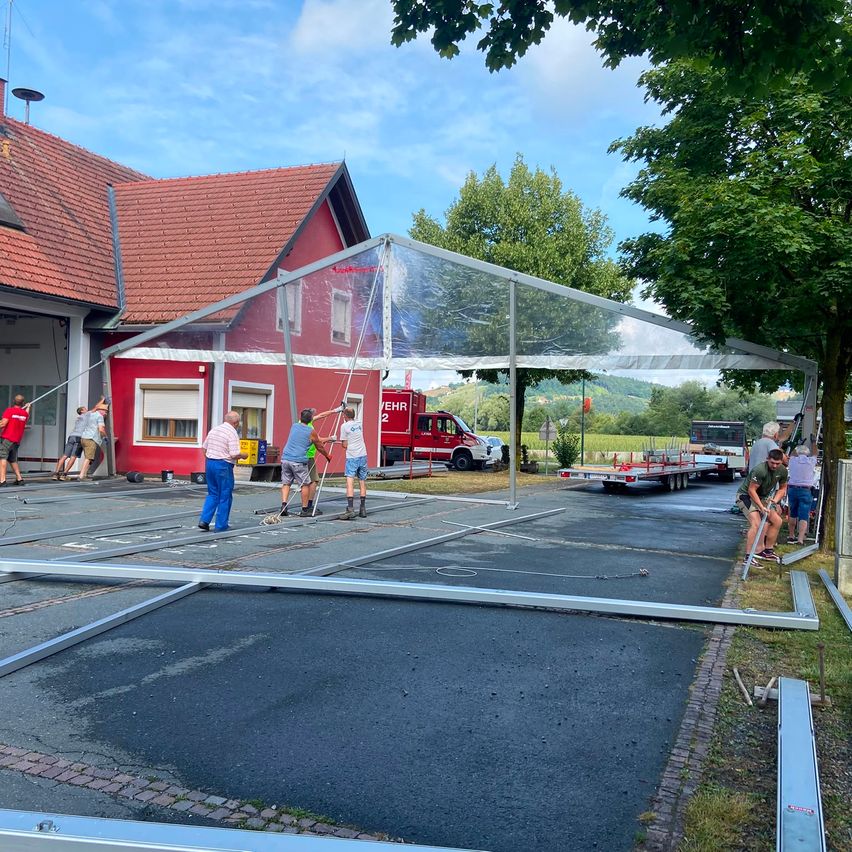 Eine Gruppe von Menschen richtet ein großes transparentes Zelt vor einem roten Gebäude auf, in dessen Nähe ein roter LKW geparkt ist.