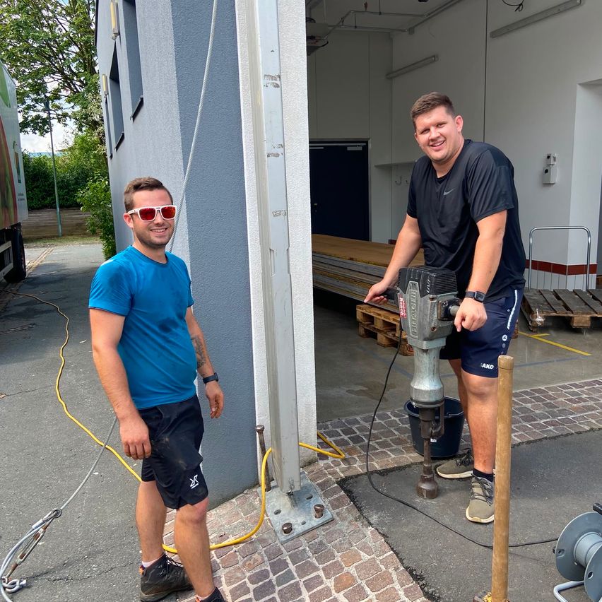 Zwei Männer stehen vor einem Gebäude mit einem Bohrgerät. Einer trägt eine Sonnenbrille und ein blaues T-Shirt, der andere ein schwarzes T-Shirt und Shorts. Sie lächeln und scheinen für ein Foto zu posieren.