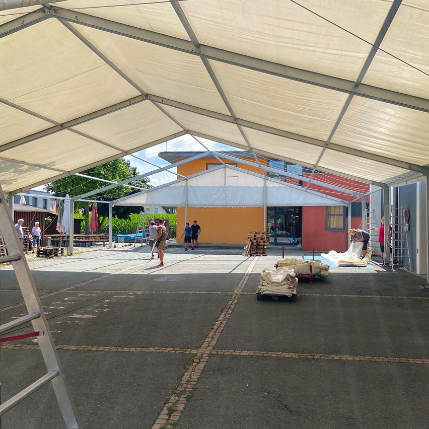 Ein großes Zelt wird in einem Außenbereich aufgebaut. Mehrere Personen arbeiten daran, einige stehen und andere auf Leitern. In der Nähe befinden sich Tische, Stühle und ein Gebäude.