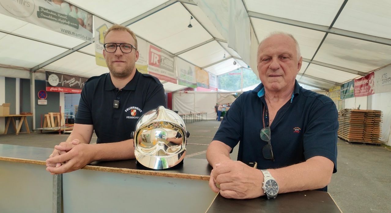 Zwei Männer in Marineblau-Poloshirts sitzen an einem Tisch mit einem glänzenden silbernen Helm vor ihnen. Der Mann auf der linken Seite trägt eine Brille und einen Namensschild. Der Mann auf der rechten Seite hat eine Uhr und eine Sonnenbrille um den Hals.