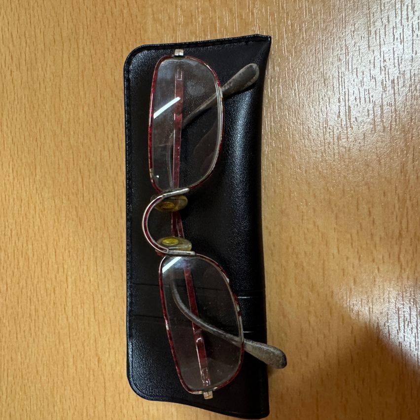 Ein Paar roter Brille mit klaren Gläsern liegt in einem schwarzen Lederetui auf einer Holzoberfläche.