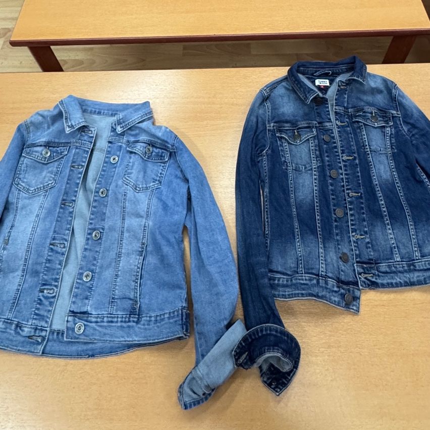 Zwei Jeansjacken, eine gefaltet und die andere flach, auf einem Holztisch. Die Jacke auf der linken Seite ist zur Hälfte gefaltet, während die Jacke auf der rechten Seite die Ärmel hochgekrempelt hat.