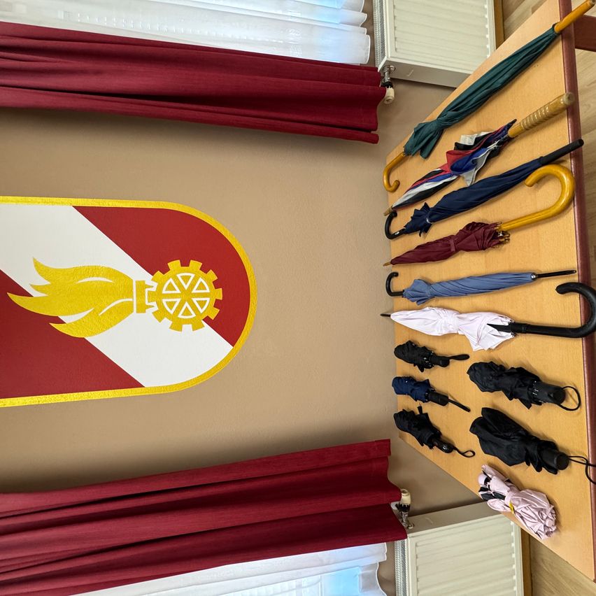 Eine Sammlung von Regenschirmen ist auf einem Holzbrett ausgestellt, mit einem roten und weißen Emblem, das eine gelbe Flamme und ein Zahnrad zeigt, an der Wand dahinter.