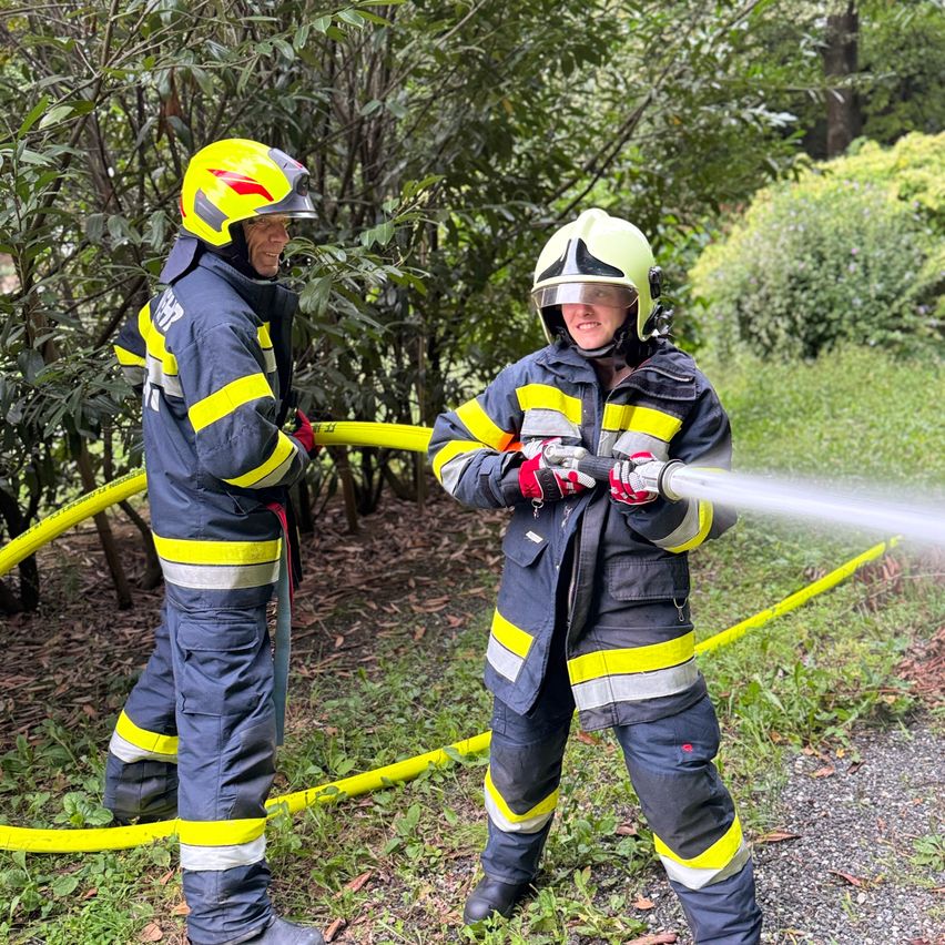 Two firefighters in full gear are spraying water from a hose in a grassy area surrounded by bushes.