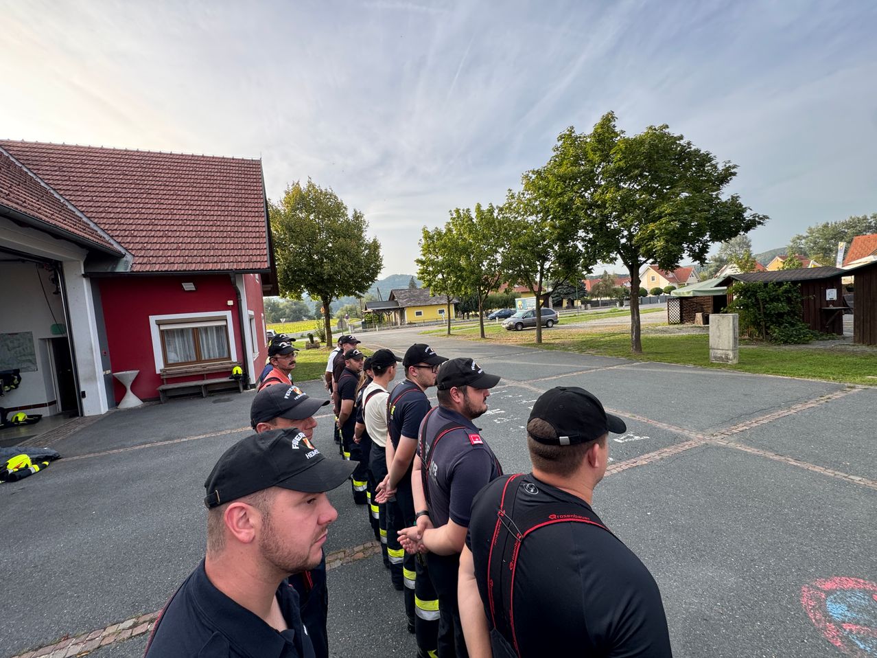 A group of firefighters in uniform stands in a line in front of a red building with a bench and trees.