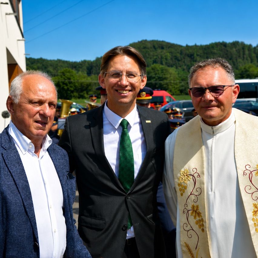 Drei Männer stehen an einem sonnigen Tag nebeneinander. Der Mann in der Mitte lächelt, während der Mann auf der rechten Seite eine Soutane und eine Sonnenbrille trägt. Hinter ihnen steht ein Gebäude, und im Hintergrund befinden sich Berge.