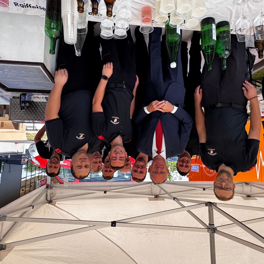 Gruppe von Menschen unter einem Zelt, Köpfe verkehrt herum, tragen schwarze Hemden und Krawatten, eine Brille auf einem Mann. Leere Flaschen und Gläser auf dem Tisch darüber.