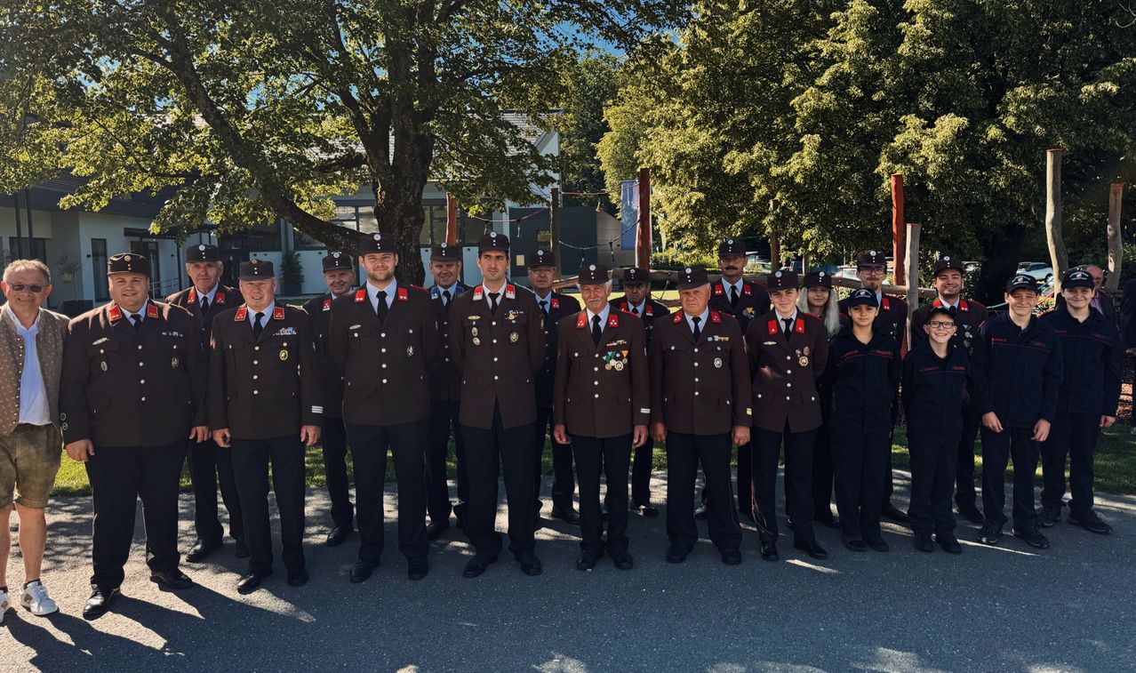 Eine Gruppe uniformierter Personen, möglicherweise Militär, steht in Formation unter einem Baum mit Gebäuden im Hintergrund.