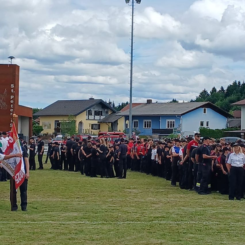 Eine große Gruppe uniformierter Personen steht auf einem Feld. Eine Person hält eine Fahne im Vordergrund. Im Hintergrund sind Häuser und ein Mast zu sehen.