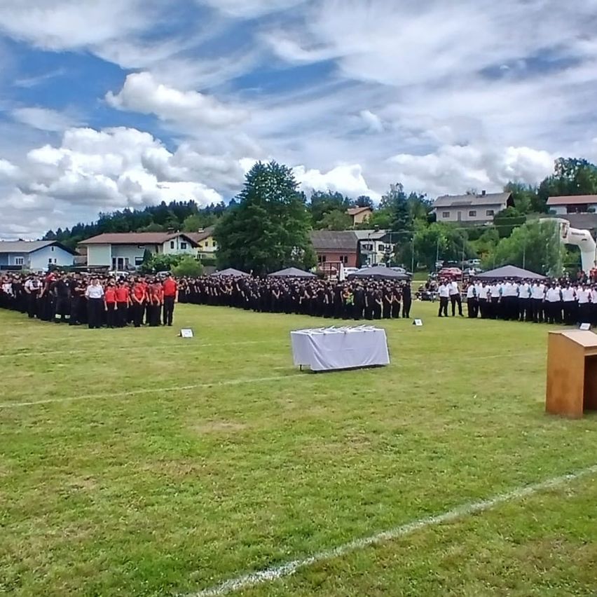 Eine große Gruppe von Menschen in roten und schwarzen Uniformen steht in Reihen auf einem Rasenfeld unter einem bewölkten Himmel. Ein Podium mit einem Tisch ist in der Mitte aufgestellt.