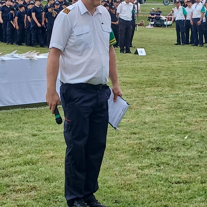 Eine Gruppe uniformierter Personen, wahrscheinlich Militärangehörige, steht in Formation auf einem Rasenfeld. Ein Mann vorne hält ein Mikrofon und ein Stück Papier.