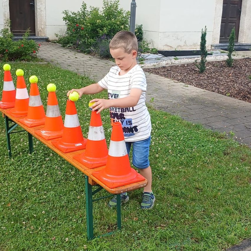 Ein Junge spielt mit Tennisbällen und orangefarbenen Verkehrskegel, die in einer Reihe auf einem Tisch im Hof angeordnet sind.