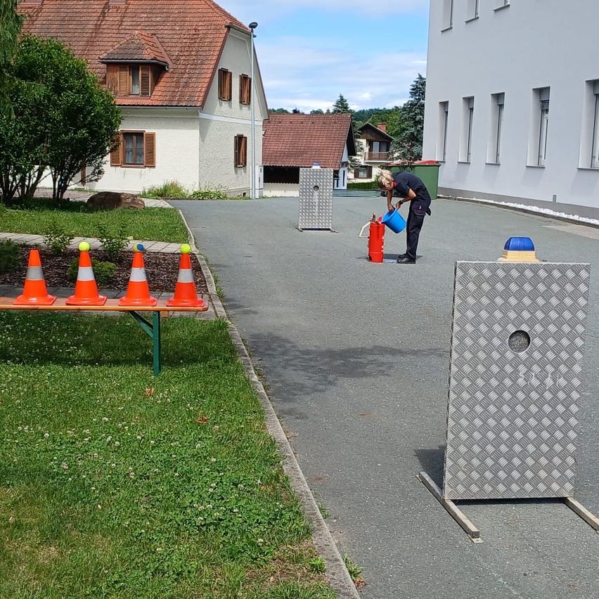 Eine Person füllt einen roten Feuerlöscher, während sie auf einem asphaltierten Weg steht, in der Nähe mehrerer Verkehrskegel und eines Metallschildes. Im Hintergrund befinden sich Häuser und Bäume.