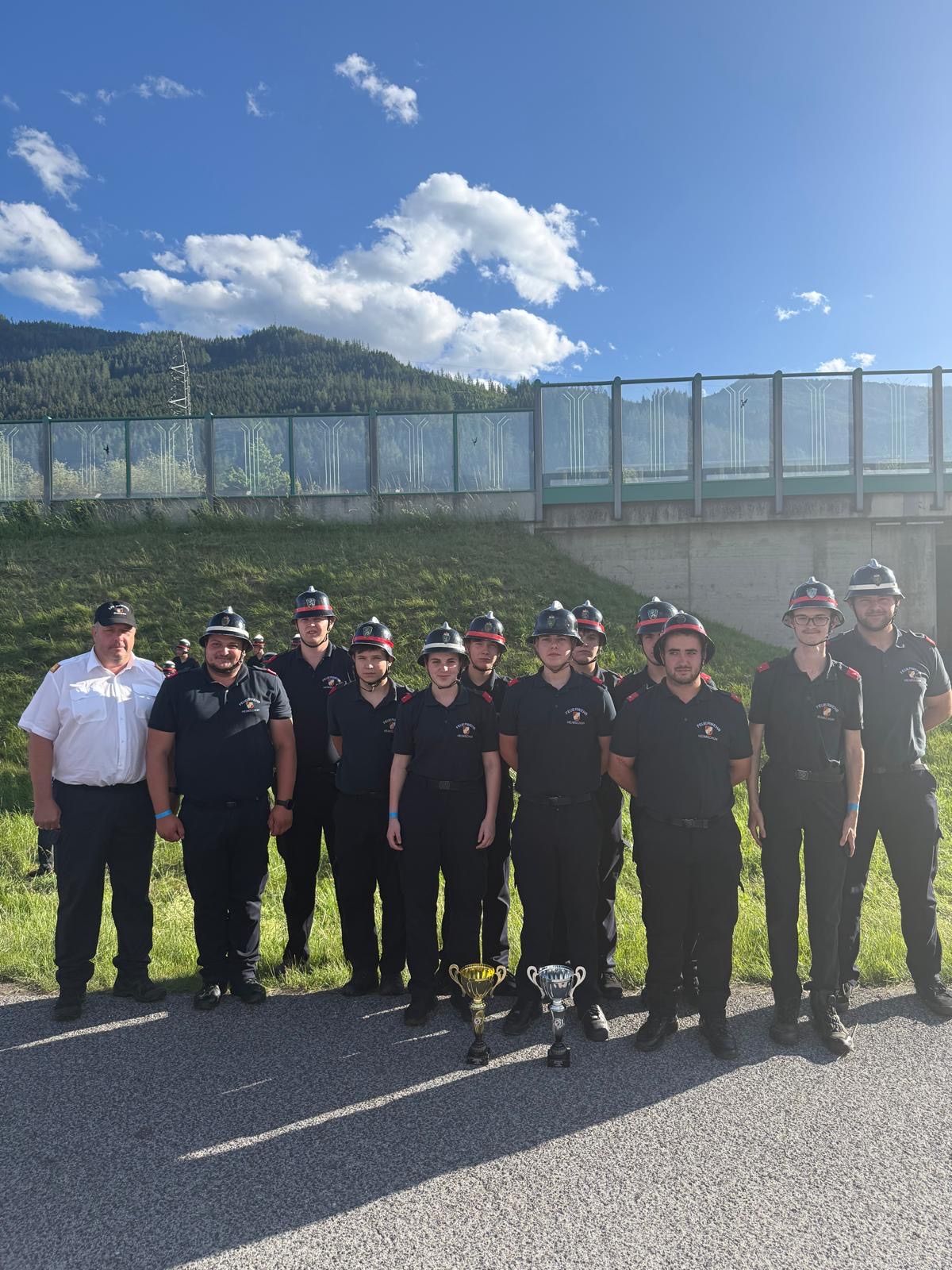 Eine Gruppe von Feuerwehrleuten in Uniform, die im Freien stehen und für ein Foto mit zwei Trophäen posieren, unter einem klaren blauen Himmel mit Bergen im Hintergrund.