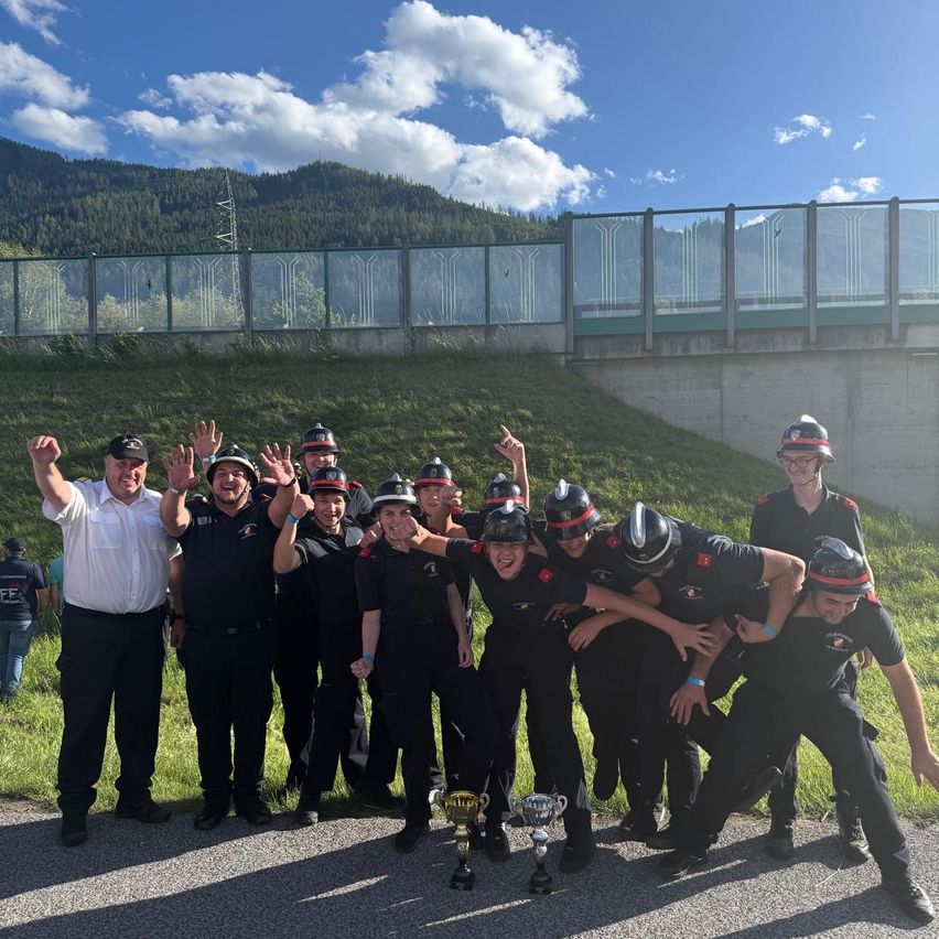 Eine Gruppe Feuerwehrleute steht mit Trophäen vor einer Brücke, mit Bergen und einem bewölkten Himmel im Hintergrund.