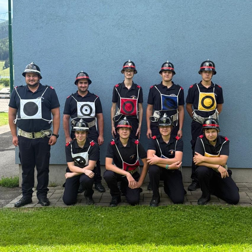 Eine Gruppe von Feuerwehrleuten in Uniform posiert für ein Foto. Sie stehen und knien auf Gras. Dahinter befindet sich eine blaue Wand.