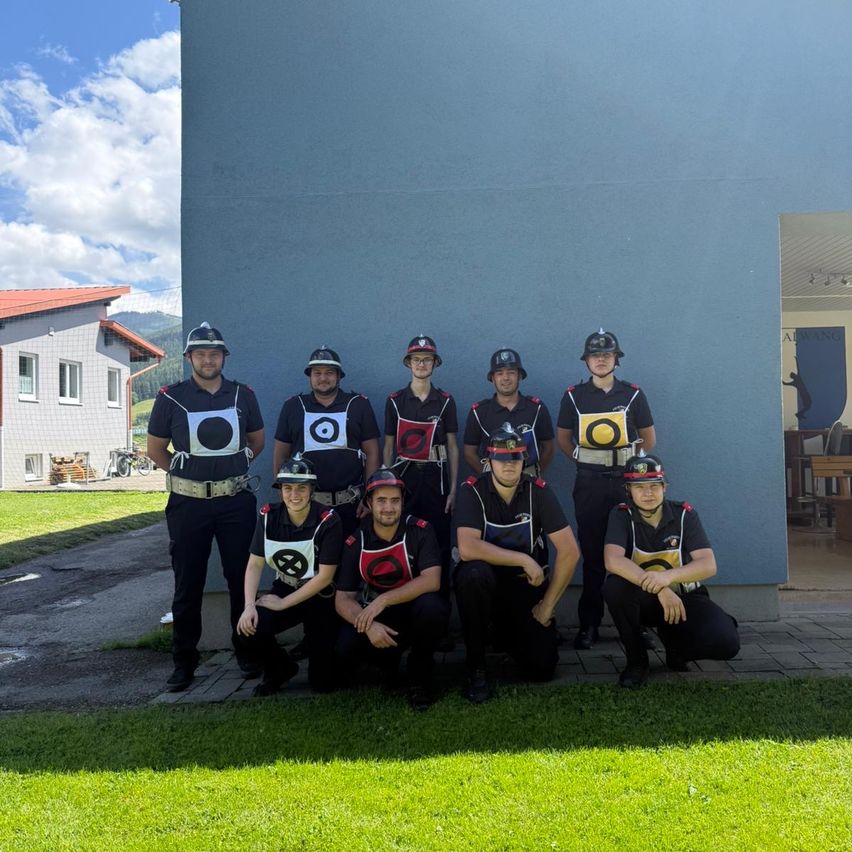 Eine Gruppe uniformierter Personen posiert für ein Foto vor einem blauen Gebäude. Sie tragen Helme mit unterschiedlich farbigen Aufnähern. Hinter ihnen ist ein Haus und ein klarer Himmel mit Wolken zu sehen.