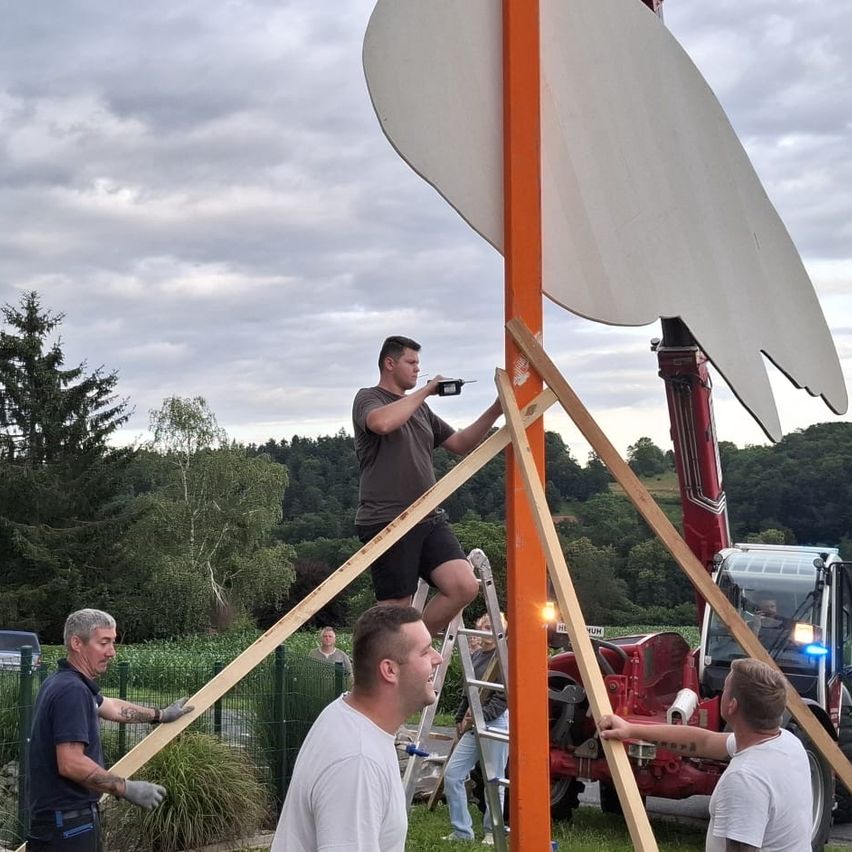Eine Gruppe von Männern arbeitet an der Errichtung einer großen Skulptur im Freien. Ein Mann klettert auf eine Leiter, ein anderer hält eine Holzstange und ein dritter steht neben einem großen roten Traktor. Bäume und ein bewölkter Himmel sind im Hintergrund.