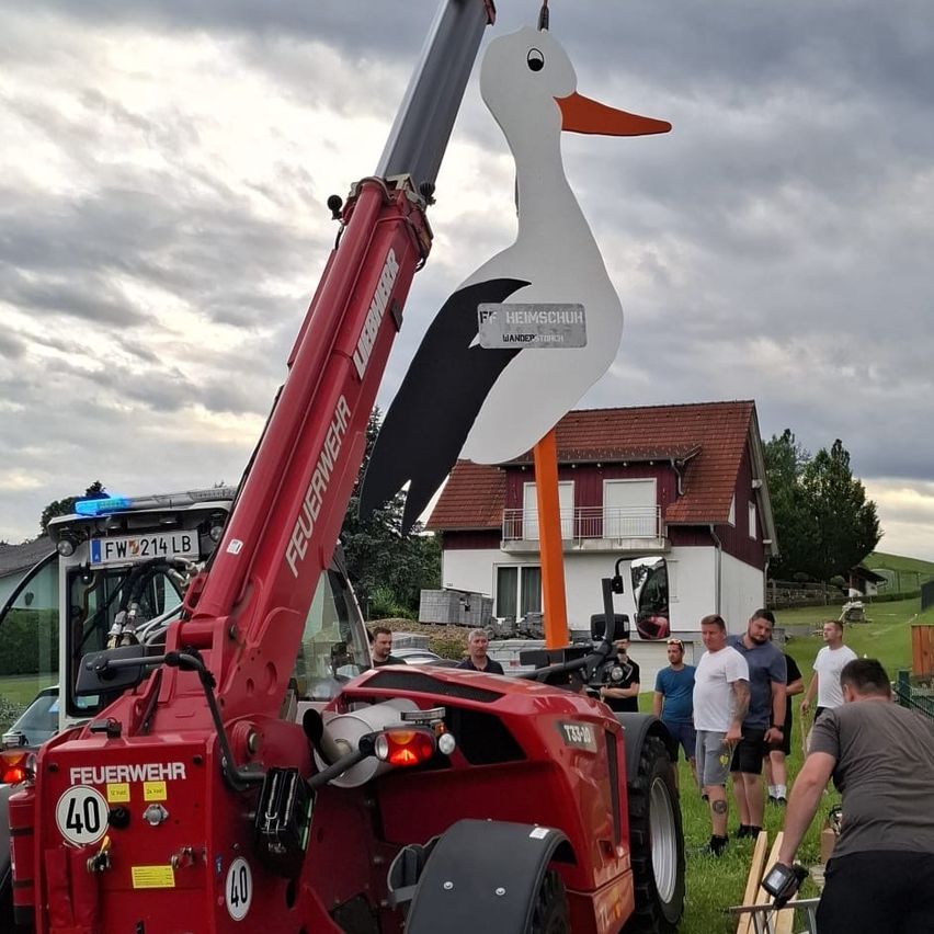 Eine große Storch-Skulptur wird von einem roten Kran vor einem Haus gehoben. Mehrere Männer stehen umher und beobachten den Vorgang.