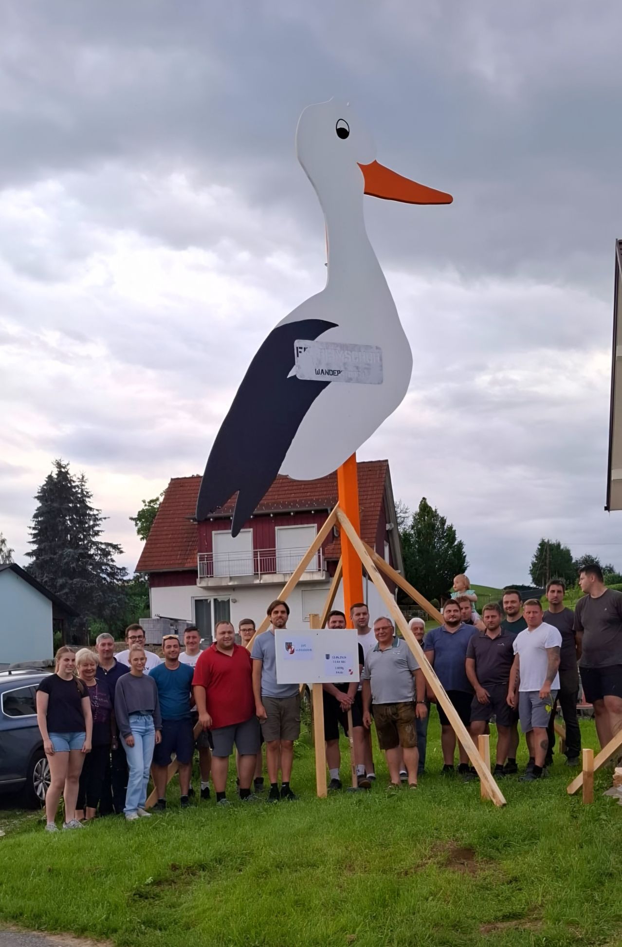Eine Gruppe von Menschen steht um eine große Storchendkulptur vor einem Haus. Die Statue besteht aus Holz und Metall.
