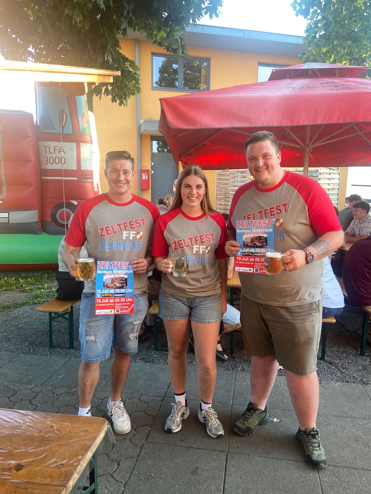 Drei Personen, zwei Männer und eine Frau, stehen zusammen draußen. Sie halten Getränke und tragen passende T-Shirts mit der Aufschrift Zeltfest. Ein roter Regenschirm und ein aufblasbarer Feuerwehrwagen sind im Hintergrund zu sehen.