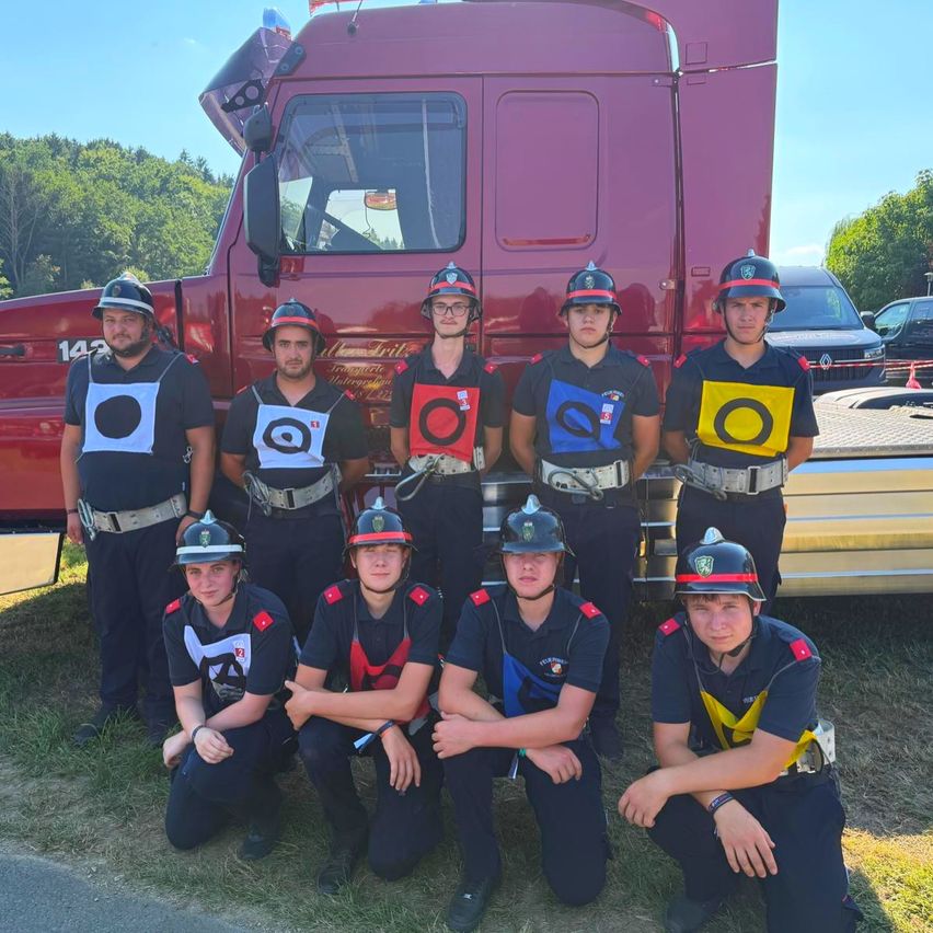 Eine Gruppe von Feuerwehrleuten posiert für ein Foto vor einem roten Lkw an einem sonnigen Tag, mit Bäumen und einem Auto im Hintergrund.