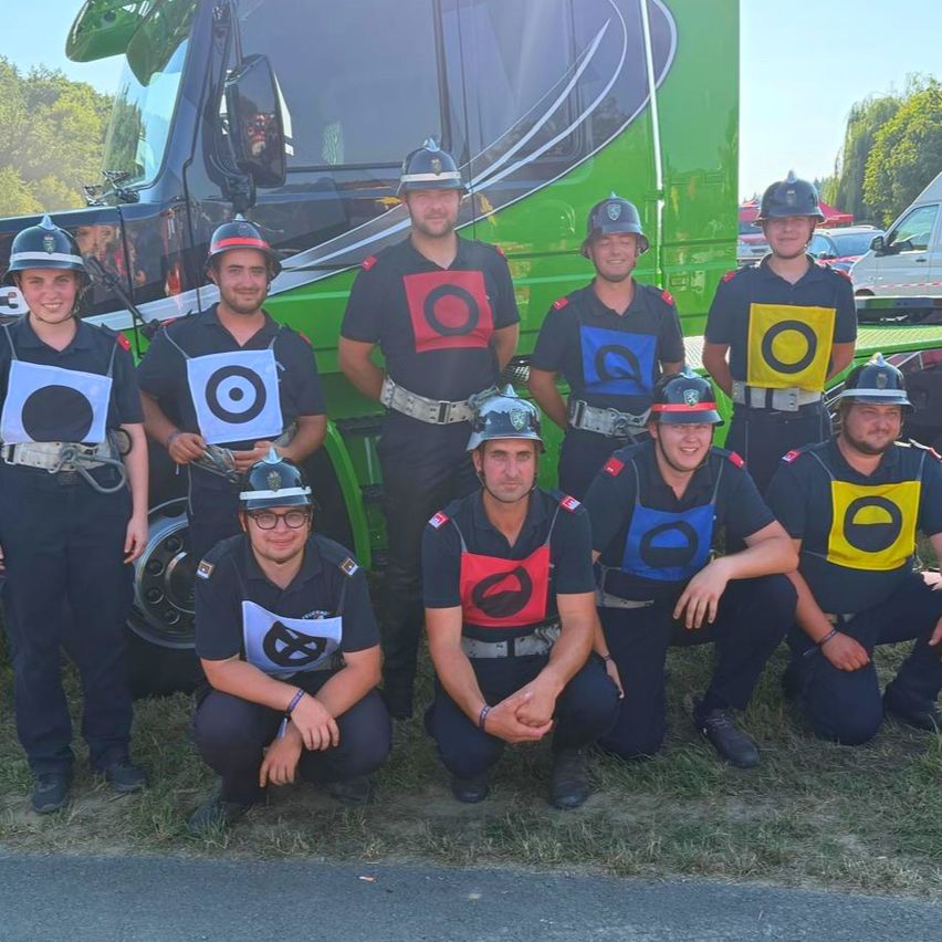 Eine Gruppe von Feuerwehrleuten in Uniform mit Helmen und Brillen posiert für ein Foto vor einem grünen Lastwagen.