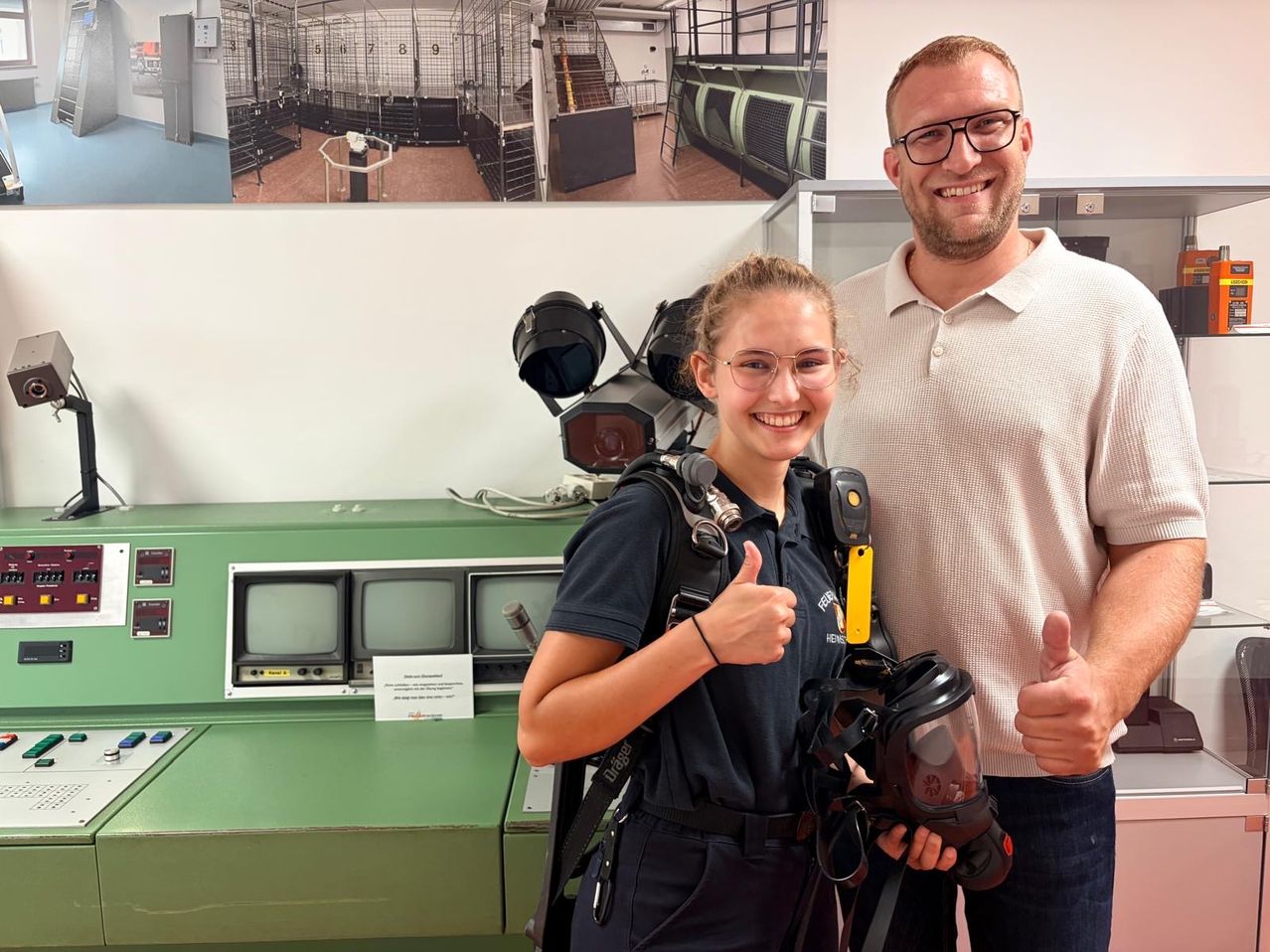 Ein Mann und eine Frau in Uniformen stehen neben einem grünen Bedienfeld, lächeln und geben Daumen hoch. Dahinter ist ein Foto von einer Treppe und Käfigen zu sehen.