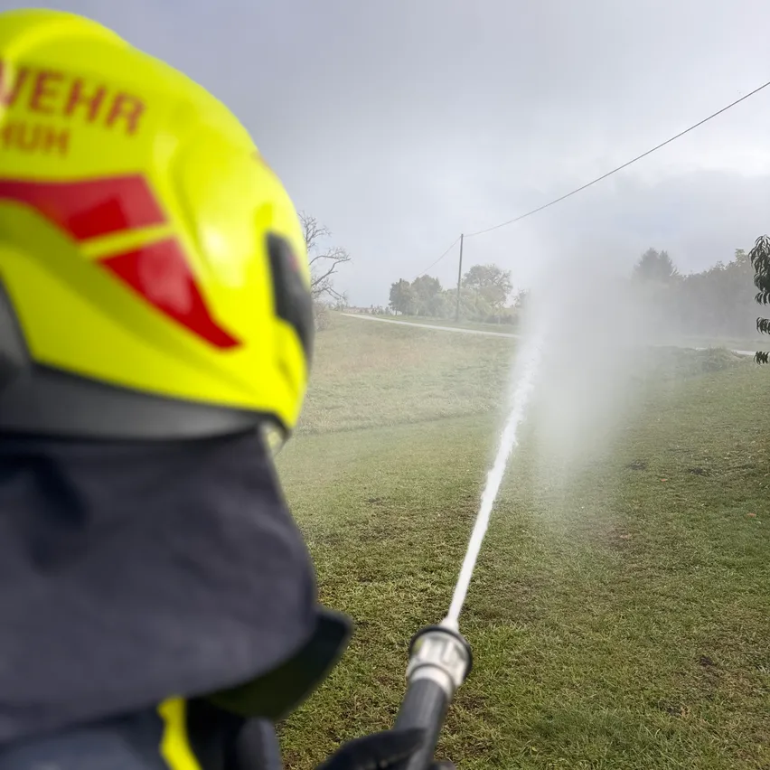 Ein Feuerwehrmann mit gelbem Helm und gelber und schwarzer Uniform sprüht Wasser aus einem Schlauch auf einer Wiese.