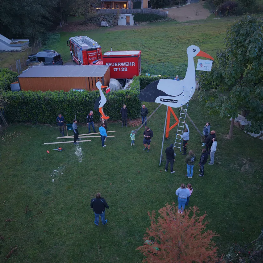 Ein Luftbild von einer Gruppe von Menschen, die in einem Grasfeld stehen. Sie sind um eine große Storchstatue versammelt, wobei einige Personen eine Leiter zur Spitze erklimmen. Im Hintergrund befindet sich ein roter Feuerwehrwagen und ein Gebäude.