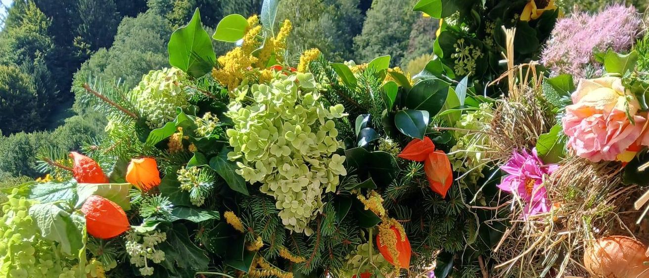 Eine kreisförmige Blumendekoration mit einer Vielzahl von Blumen, Blättern und Gemüse, einschließlich Kürbisse, Karotten und Trauben. Sie ist im Freien auf einer Holzoberfläche ausgestellt.